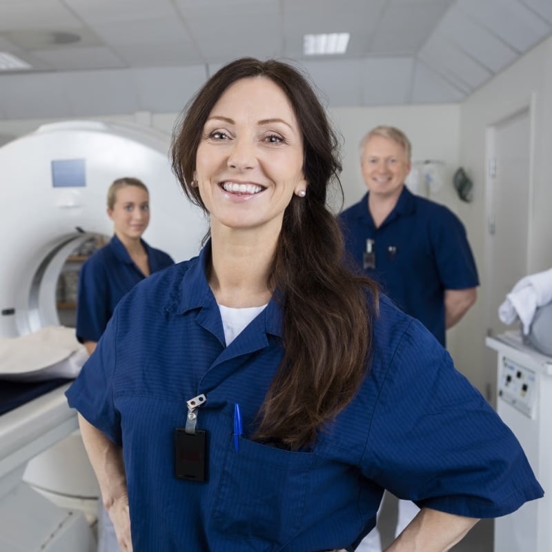 A smiling technologist in blue scrubs stands in the foreground while two colleagues stand behind near a CT scanner.